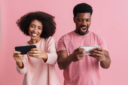 Foto de Negro feliz hombre y mujer sonriendo y utilizando el teléfono celular juntos aislados sobre fondo rosa - Imagen libre de derechos