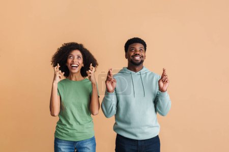 Téléchargez les photos : Noir homme et femme exprimant le bonheur et debout avec les doigts croisés isolé sur fond beige - en image libre de droit