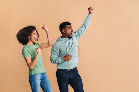 Foto de Negro hombre y mujer riendo y haciendo gestos como ganadores aislados sobre fondo beige - Imagen libre de derechos