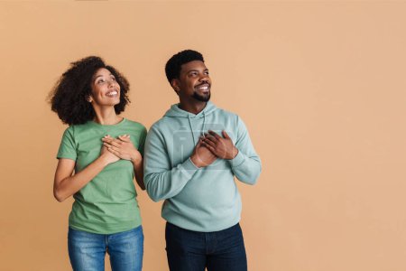 Téléchargez les photos : Homme et femme noirs exprimant leur dignité et gardant leurs mains au cœur isolé sur fond beige - en image libre de droit