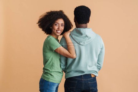 Téléchargez les photos : Femme noire exprimant le bonheur tout en embrassant l'homme de son dos isolé sur fond beige - en image libre de droit