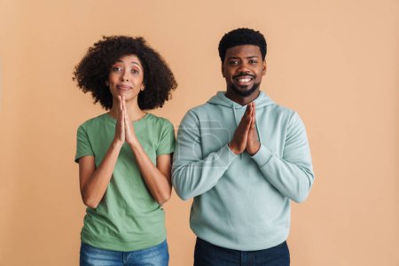 Foto de Negro hombre y mujer expresando felicidad y orando con las manos juntas aisladas sobre fondo beige - Imagen libre de derechos