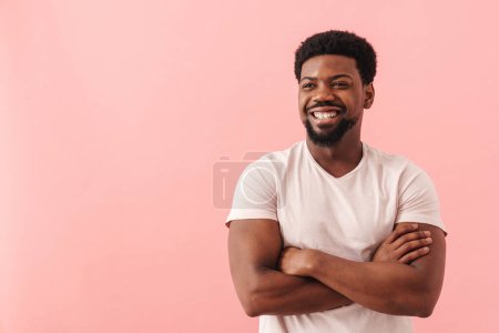 Téléchargez les photos : Noir mi-homme souriant tout en posant avec les bras croisés isolé sur fond rose - en image libre de droit
