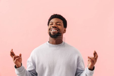 Téléchargez les photos : Noir mi-homme souriant et tenant les doigts croisés pour bonne chance isolé sur fond rose - en image libre de droit
