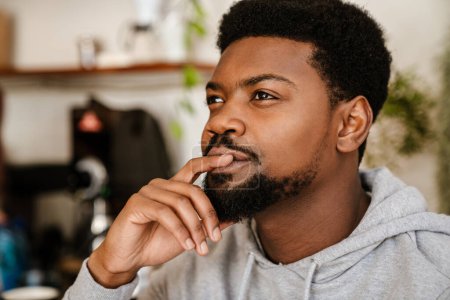 Téléchargez les photos : Homme barbu noir portant un sweat à capuche regardant de côté tout en étant assis dans un café à l'intérieur - en image libre de droit