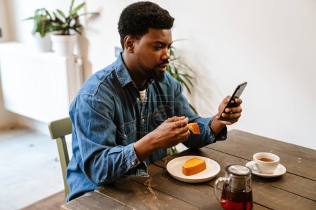 Téléchargez les photos : Homme barbu noir utilisant un téléphone portable tout en mangeant du gâteau dans un café à l'intérieur - en image libre de droit