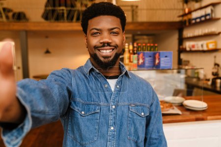 Téléchargez les photos : Noir barbu homme souriant tout en prenant selfie photo dans le café à l'intérieur - en image libre de droit