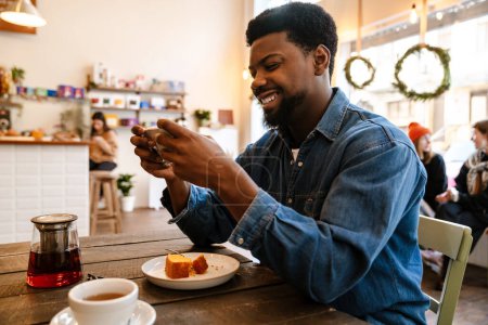 Téléchargez les photos : Homme souriant noir utilisant un téléphone portable tout en ayant brunch dans un café à l'intérieur - en image libre de droit
