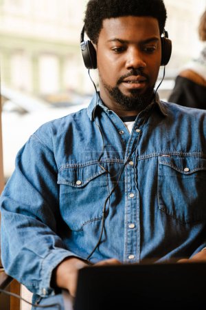 Téléchargez les photos : Noir barbu homme écouter de la musique tout en utilisant un ordinateur portable dans le café à l'intérieur - en image libre de droit