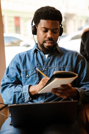 Téléchargez les photos : Homme barbu noir note tout en utilisant un ordinateur portable dans le café à l'intérieur - en image libre de droit