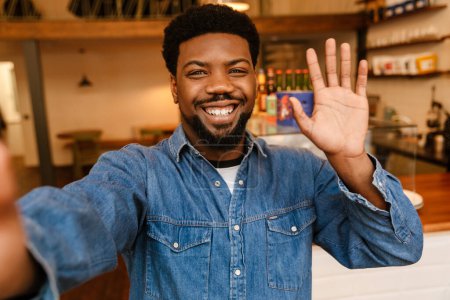 Photo for Black bearded man waving hand while taking selfie photo in cafe indoors - Royalty Free Image