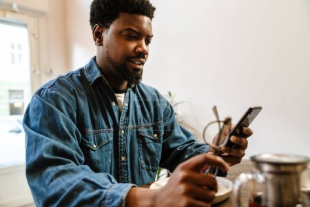 Téléchargez les photos : Homme barbu noir utilisant un téléphone portable tout en étant assis dans un café à l'intérieur - en image libre de droit