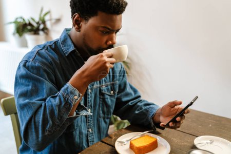 Téléchargez les photos : Homme barbu noir utilisant un téléphone portable tout en buvant du thé dans un café à l'intérieur - en image libre de droit