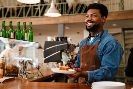 Téléchargez les photos : Homme barbu noir portant tablier riant tout en travaillant dans un café à l'intérieur - en image libre de droit
