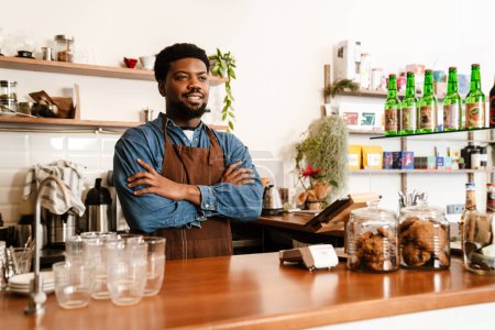 Téléchargez les photos : Homme barbu noir portant tablier souriant tout en travaillant dans un café à l'intérieur - en image libre de droit