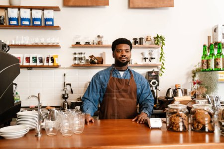Téléchargez les photos : Homme barbu noir portant tablier souriant tout en travaillant dans un café à l'intérieur - en image libre de droit