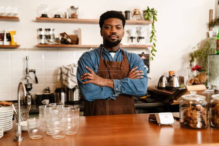 Téléchargez les photos : Homme barbu noir portant tablier debout avec les bras croisés dans le café à l'intérieur - en image libre de droit