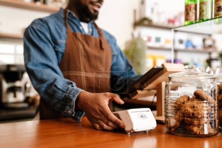 Téléchargez les photos : Noir barbu homme portant tablier de travail avec caisse enregistreuse dans le café à l'intérieur - en image libre de droit