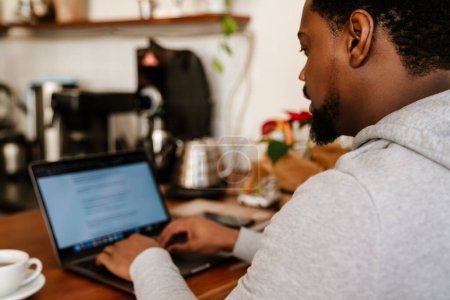 Téléchargez les photos : Homme barbu noir tapant sur ordinateur portable tout en travaillant dans un café à l'intérieur - en image libre de droit