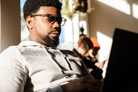 Foto de Negro barbudo hombre trabajando con el ordenador portátil mientras está sentado en la cafetería en el interior - Imagen libre de derechos