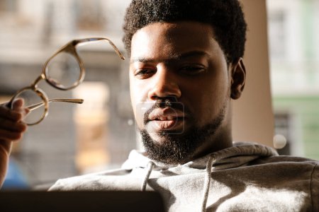 Téléchargez les photos : Noir barbu homme tenant des lunettes tout en travaillant avec ordinateur portable dans le café à l'intérieur - en image libre de droit