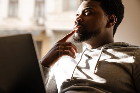 Téléchargez les photos : Noir perplexe homme travaillant avec ordinateur portable tout en étant assis dans le café à l'intérieur - en image libre de droit