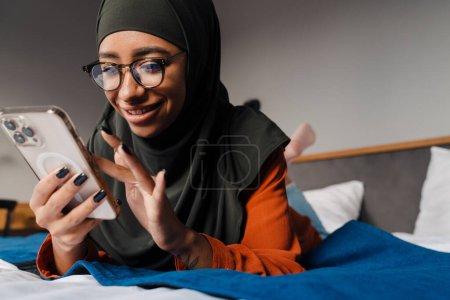 Foto de Joven hermosa mujer sonriente en hijab y gafas acostada en la cama y usando su teléfono - Imagen libre de derechos