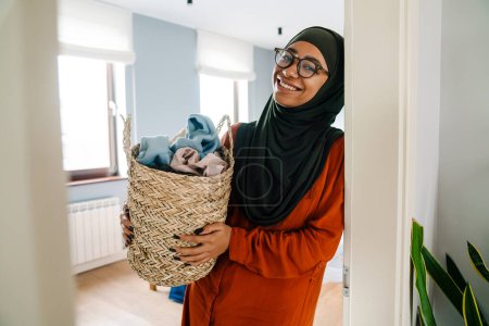 Téléchargez les photos : Jeune belle femme souriante en hijab et des lunettes tenant panier loundry et regardant la caméra dans la chambre ensoleillée à la maison - en image libre de droit
