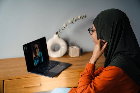 Téléchargez les photos : Jeune femme en hijab et lunettes tenant une vidéoconférence assis dans une chambre confortable à la maison - en image libre de droit