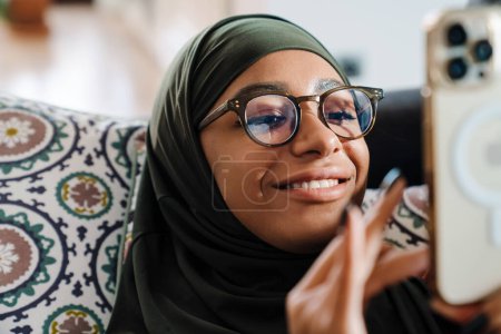 Foto de Joven hermosa mujer sonriente en hijab y gafas mirando en su teléfono - Imagen libre de derechos