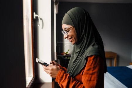 Foto de Joven hermosa mujer sonriente en hijab y gafas mirando en su teléfono delante de la ventana en casa - Imagen libre de derechos
