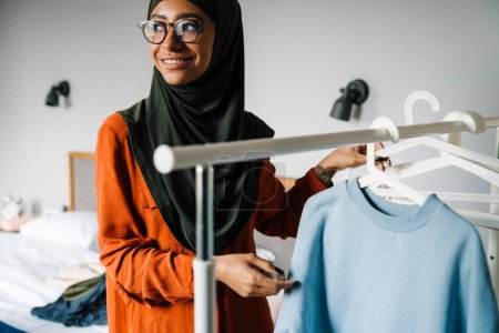 Photo for Young beautiful smiling woman in hijab and glasses hanging clothes on rail at home - Royalty Free Image