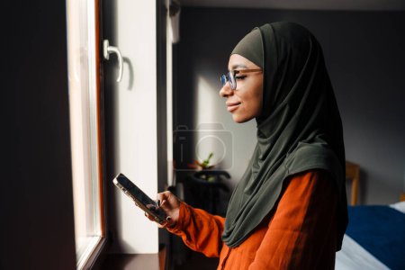 Foto de Joven hermosa mujer tranquila en hijab y gafas con teléfono delante de la ventana en casa - Imagen libre de derechos