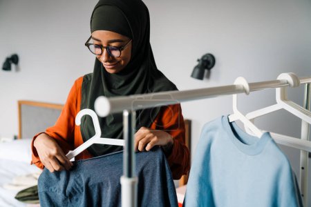 Téléchargez les photos : Jeune belle femme souriante en hijab et des lunettes accrochant des vêtements sur cintre à la maison - en image libre de droit