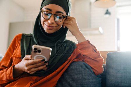 Foto de Joven hermosa mujer sonriente en hijab y gafas sentadas en el sofá con el teléfono y apuntalando su cabeza - Imagen libre de derechos