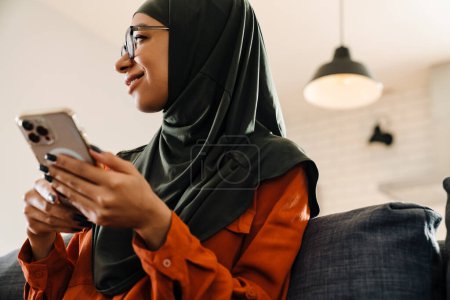 Téléchargez les photos : Jeune belle femme souriante en hijab et des lunettes assis sur le canapé avec téléphone et regardant vers la gauche - en image libre de droit