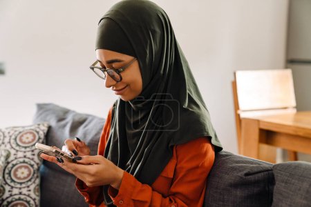 Foto de Joven hermosa mujer sonriente en hijab y gafas sentadas en el sofá y mirando en su teléfono - Imagen libre de derechos