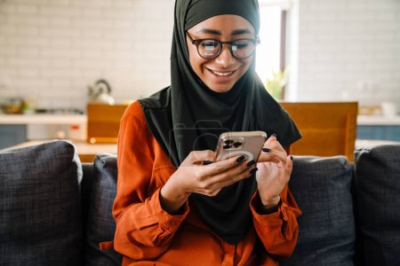 Foto de Joven hermosa mujer sonriente en hijab y gafas sentadas en el sofá y mirando en su teléfono - Imagen libre de derechos