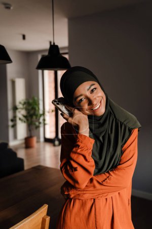 Téléchargez les photos : Jeune belle femme souriante heureuse dans le hijab avec le téléphone soutenant sa tête et regardant la caméra - en image libre de droit
