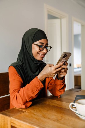 Téléchargez les photos : Jeune belle femme souriante en hijab et des lunettes assises à la table et regardant son téléphone - en image libre de droit