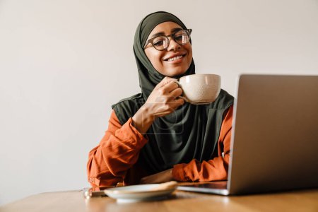 Foto de Joven hermosa mujer sonriente en hijab y vasos sosteniendo la taza de té mientras trabaja con el ordenador portátil y mirando a la cámara - Imagen libre de derechos