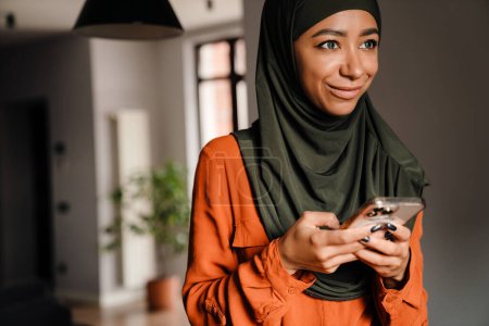 Téléchargez les photos : Jeune belle femme souriante en hijab avec téléphone debout à la maison - en image libre de droit