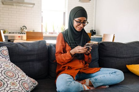 Téléchargez les photos : Jeune belle femme souriante en hijab et des lunettes assises sur un canapé en pose de lotus et regardant son téléphone - en image libre de droit