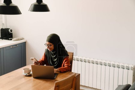 Téléchargez les photos : Jeune belle femme calme dans le hijab et des lunettes regardant sur son téléphone tout en travaillant avec ordinateur portable dans une cuisine confortable à la maison - en image libre de droit