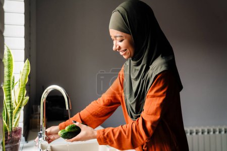 Foto de Joven feliz sonriente mujer en hijab lavando pepinos para preparar ensalada en casa - Imagen libre de derechos
