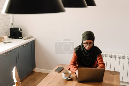 Foto de Joven hermosa mujer tranquila en hijab y gafas de trabajo con el ordenador portátil en la acogedora cocina en casa - Imagen libre de derechos