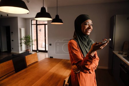 Foto de Joven hermosa mujer sonriente feliz en hijab con teléfono mirando hacia la derecha mientras está de pie en la acogedora cocina en casa - Imagen libre de derechos