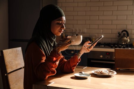Téléchargez les photos : Vue de côté de jeune belle femme calme en hijab assis à la table de cuisine avec tasse de biscuits de thé et téléphone - en image libre de droit