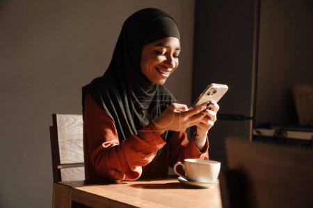Téléchargez les photos : Jeune belle femme souriante en hijab assise à la table de cuisine avec une tasse de thé et regardant son téléphone - en image libre de droit