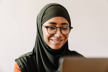 Foto de Joven hermosa mujer sonriente en hijab y gafas de trabajo con el primer plano portátil - Imagen libre de derechos